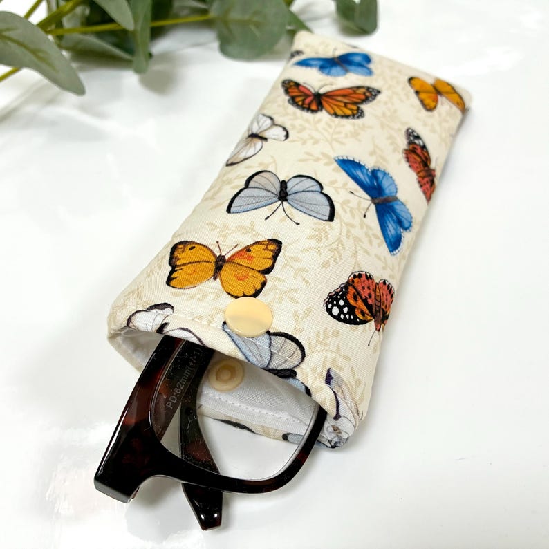 May include: A beige eyeglasses case with a colorful butterfly pattern. The case is open, revealing a pair of eyeglasses. The case has a button closure and is made of fabric.