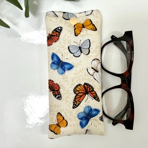 May include: A cream-colored glasses case with a button closure, adorned with colorful butterfly patterns. Next to the case are a pair of tortoiseshell eyeglasses. The butterflies are in shades of orange, blue, red, and white.