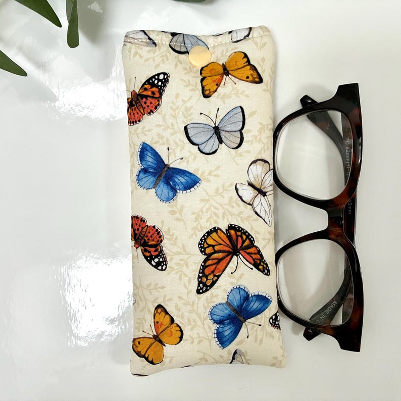 May include: A cream-colored glasses case with a button closure, adorned with colorful butterfly patterns. Next to the case are a pair of tortoiseshell eyeglasses. The butterflies are in shades of orange, blue, red, and white.