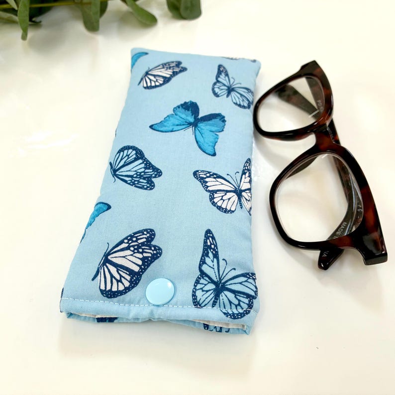 May include: A light blue glasses case with a pattern of blue and white butterflies. The case is rectangular with a light blue button closure. Tortoiseshell eyeglasses are next to the case.