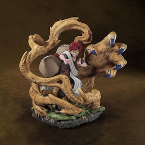 May include: A detailed figurine of a character with red hair and a white scarf, surrounded by a sand-like structure. The figure is posed dynamically, with a determined expression. The base is a mix of green and grey.