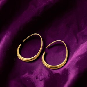 May include: A pair of gold-toned hoop earrings with an irregular, organic oval shape. The earrings feature a smooth, polished metallic finish and a continuous, open-ended design that curves into a rounded silhouette, creating a simple, sleek look.