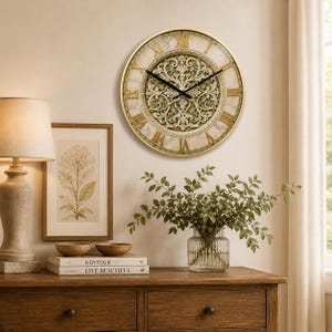 May include: A circular wall clock with a gold-coloured frame and Roman numerals. The clock face has a marble-effect background with an ornate, carved design in the centre. Black clock hands show the time. The clock is mounted on a beige wall.