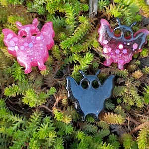 Può includere: Tre ornamenti in resina a forma di pipistrello. Uno è nero, uno rosa con glitter e l'altro è rosa scuro con glitter. Gli ornamenti sono su un letto di piante verdi.