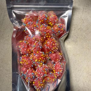 May include: A transparent, sealed bag containing numerous pink, spherical sweets coated in colourful sprinkles. The candies have a textured surface and are packed together. A small packet with the text "DO NOT EAT" is visible inside the bag.