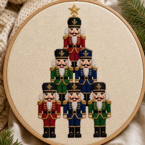 May include: A cross-stitch embroidery in a circular wooden hoop featuring six nutcracker figures arranged in a pyramid shape. The nutcrackers wear red, green, and blue uniforms with black hats and gold accents, topped with a single gold star.