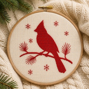 May include: A circular wooden embroidery hoop containing a cross-stitch pattern on off-white fabric. The design features a solid red cardinal bird perched on a pine branch, surrounded by small red stitched snowflakes against the fabric background.