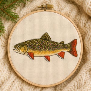 May include: A circular wooden embroidery hoop containing a cross-stitch design of a brook trout. The fish features a dark olive green back with small red and blue spots, a yellow-toned belly, and vibrant orange-red fins on a cream fabric.