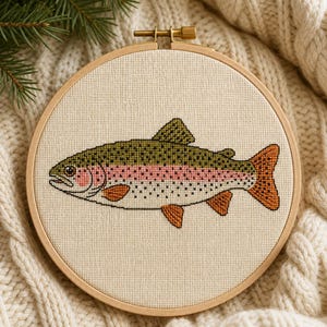 May include: A circular wooden embroidery hoop containing a cross-stitch design of a rainbow trout. The fish features a green back with dark spots, a pink lateral stripe, and orange fins, all stitched onto a light beige fabric background.