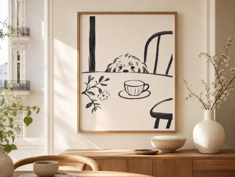 May include: A framed art print featuring a whimsical illustration of a dog peeking over a table. The black and white drawing includes a teacup, chair, and floral accents. The artwork is displayed in a room with a wooden cabinet and a white vase.