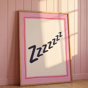 以下が含まれることがあります： ライトベージュの背景にネイビーブルーの「Zzzzzz」の文字が入ったフレーム入りプリント。ピンクの縁取りと内側の細い赤い線があり、明るい木製のフレームに入っています。アートワークはピンクの壁に展示されています。
