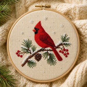 May include: A cross-stitch embroidery in a round wooden hoop featuring a bright red cardinal bird perched on a snow-covered pine branch with green needles, a brown pinecone, and clusters of red berries against a cream-colored fabric background.