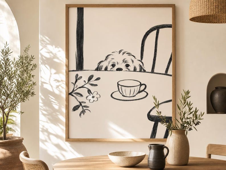 May include: A framed black and white illustration of a dog peeking over a table with a teacup and saucer. The artwork is in a wooden frame and is displayed in a room with a table, vase, and other decor.