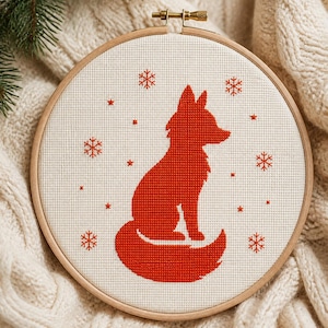 May include: A circular wooden embroidery hoop containing a piece of white fabric with a cross-stitch design. The design features a solid red fox silhouette surrounded by several small red embroidered snowflakes and tiny star-shaped accents.