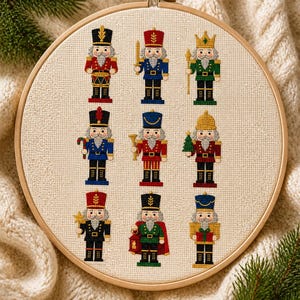 Puede incluir: Un bastidor de bordado circular de madera que contiene un patrón de punto de cruz de nueve figuras de cascanueces dispuestas en tres filas. Cada cascanueces viste un uniforme distinto en colores rojo, azul, verde, negro y dorado.