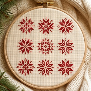May include: A circular wooden embroidery hoop containing a piece of cream-colored Aida fabric. The fabric features nine distinct, symmetrical snowflake patterns stitched in red thread, arranged in a three-by-three grid formation.