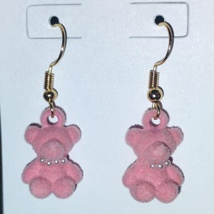 May include: A pair of pink teddy bear earrings. Each earring features a fuzzy, pink teddy bear charm with a pearl necklace. The earrings have gold-colored fish hook ear wires and are displayed on a white card.