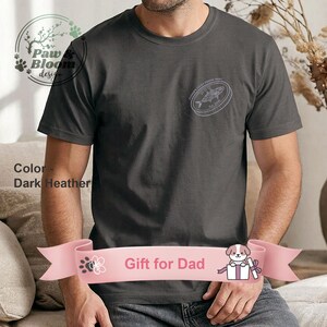 Puede incluir: Camiseta gris jaspeado oscuro con un gráfico circular en el pecho que dice "Father's Lucky Fishing Buddy" con una ilustración de un pez. La camiseta también tiene el texto "Gift for Dad" en una pancarta rosa.