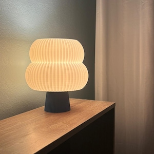 Peut inclure: Une lampe de table moderne avec un abat-jour côtelé blanc cassé et une base bleu foncé. La lampe est allumée, dégageant une lueur chaleureuse. Elle est posée sur une table en bois, avec un mur vert foncé et un rideau blanc en arrière-plan.