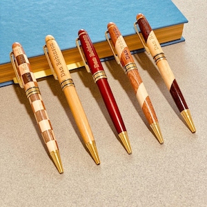 Pode incluir: Cinco canetas de madeira artesanais com detalhes e pontas douradas. Cada caneta apresenta um padrão de grão de madeira único, incluindo um design xadrez e um acabamento vermelho escuro. O texto "Dan Robinson" está gravado em cada caneta.