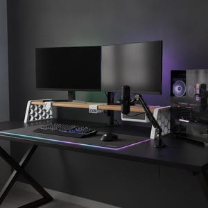 Könnte beinhalten: Ein schwarzer Schreibtisch mit zwei Monitoren auf einem Holzständer mit weißen geometrischen Stützen. Eine schwarze mechanische Tastatur und Maus liegen auf einem großen RGB-Mauspad. Ein schwarzer PC-Tower mit beleuchteten Lüftern ist vorhanden.