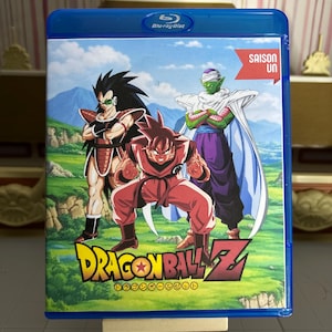 May include: A blue Blu-ray disc case with Dragon Ball Z characters on the cover. The artwork depicts three figures in dynamic poses set against a mountain and green field background. The Dragon Ball Z logo is visible.