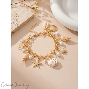 Könnte beinhalten: Goldfarbenes Charm-Armband mit verschiedenen Muschel-Anhängern, Kunstperlen und einem Seestern-Anhänger. Das Armband hat einen Karabinerverschluss und wird auf einer neutralen Oberfläche präsentiert. Der Text "Celoura Jewelry" ist sichtbar.
