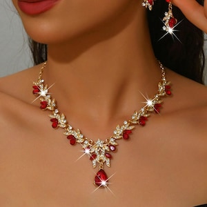 Puede incluir: Un conjunto de collar y pendientes dorados con piedras preciosas rojas y transparentes. El collar presenta un colgante en forma de lágrima y un diseño floral. Los pendientes tienen un diseño floral a juego. La joyería está sobre un fondo neutro.