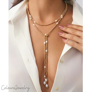 Op de afbeelding: Een goudkleurige lariat-ketting met een dubbellaags kettingontwerp bestaande uit kleine metalen kraaltjes afgewisseld met witte, ovaalvormige parels. De ketting hangt in een Y-vorm met twee bungelende strengen die eindigen in parels.