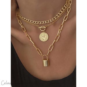 Op de afbeelding: Een goudkleurige gelaagde ketting set. De bovenste laag is een gourmetketting, gevolgd door een delicate ketting met een munt hanger en een langere ketting met een hangslot hanger. De kettingen worden op een nek getoond.