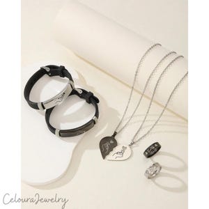 Op de afbeelding: Een collectie bijpassende sieraden, waaronder twee zwarte armbanden met zilveren accenten, een gesplitste hartvormige hangerketting en twee ringen. De armbanden en hanger hebben handgebaarontwerpen. De ringen hebben gegraveerde patronen en tekst.