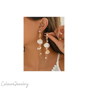 Può includere: Orecchini pendenti color oro con dettagli di conchiglie e perle. Gli orecchini hanno un ciondolo a forma di stella marina e delicate catene dorate. Il design include conchiglie bianche e dorate e piccole perle rotonde. Gli orecchini sono un gioiello a tema spiaggia.