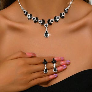 Könnte beinhalten: Ein silbernes Halsketten- und Ohrring-Set. Die Halskette hat schwarze, blattförmige Akzente und klare, funkelnde Steine. Die Ohrringe spiegeln das Design wider, mit schwarzen tropfenförmigen Steinen und klaren Akzenten. Der Schmuck wird an einer Person gezeigt.