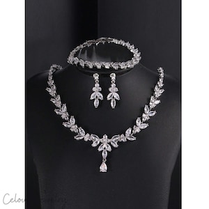 Puede incluir: Un conjunto de joyas de novia con collar, pulsera y pendientes. El conjunto es plateado y está adornado con cristales transparentes y brillantes. El collar tiene un diseño de hojas con un colgante en forma de lágrima.