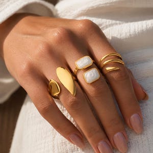 Könnte beinhalten: Ein Set aus goldfarbenen Ringen, bestehend aus einem Ring mit offenem Oval-Design, einem Ring mit zwei rechteckigen weißen Steineinlagen und einem Ring im dreifachen Wickel-Stil. Die Ringe sind poliert und metallisch mit glatten Oberflächen.