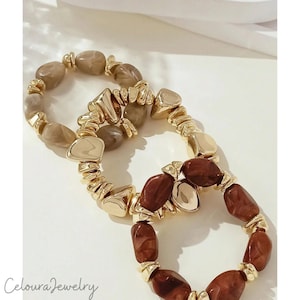Puede incluir: Dos pulseras de cuentas. Una pulsera presenta cuentas marrones claras y doradas, mientras que la otra tiene cuentas marrones oscuras y doradas. Las pulseras están dispuestas sobre una superficie blanca. La marca "Celoura Jewelry" es visible abajo a la izquierda.