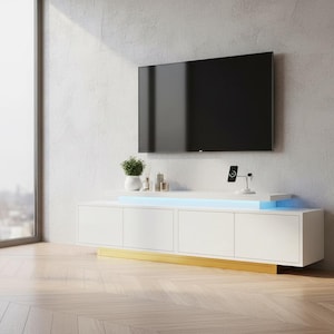 May include: A white media console with four cabinet doors and a gold-toned base. The unit features a top shelf with integrated blue LED lighting, a potted green plant, and a small wireless charging stand for a smartphone on the surface.