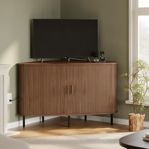 May include: A brown wooden corner media console featuring a curved front with vertical slatted detailing, two cabinet doors with slim vertical handles, and four thin black metal legs. A flat-screen television sits on the top surface.