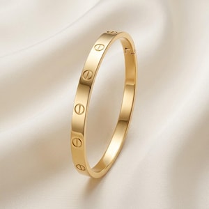 May include: A polished gold-tone bangle bracelet featuring a series of evenly spaced, engraved screw-head motifs along the outer surface. The circular band has a smooth, reflective finish and a visible hinge mechanism on one side of the structure.