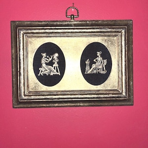 Pode incluir: Uma obra de arte emoldurada com duas silhuetas ovais pretas e figuras brancas. Uma mostra uma pessoa esculpindo um busto, a outra uma pessoa sentada. A moldura dourada tem um cabide decorativo de metal. Fundo vermelho.