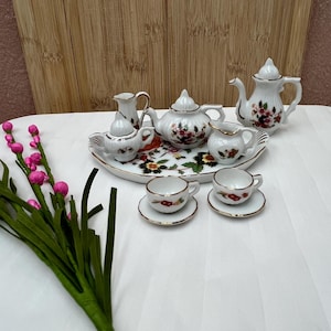 May include: A miniature porcelain tea set featuring a white base with delicate floral patterns in red, yellow, and green, accented by gold trim. The set includes a teapot, creamer, sugar bowl, two cups with saucers, and a serving tray.