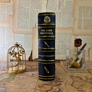 May include: A black book with gold accents and the title "HIS DARK MATERIALS" is displayed. The book has a decorative design with a knife and a telescope. The author's name, Philip Pullman, is also visible. The book is standing upright.