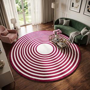 Könnte beinhalten: Ein großer runder Teppich mit einem sich wiederholenden Muster aus konzentrischen Ringen in abwechselndem tiefem Magenta und Weiß. Der Teppich liegt mittig auf einem dunklen Holzboden und bildet einen markanten geometrischen Blickfang.