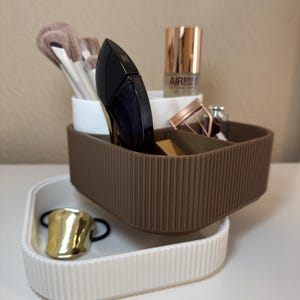 Könnte beinhalten: Ein brauner und weißer gerippter Kunststoff-Organizer mit Kosmetikprodukten. Der Organizer enthält Make-up-Pinsel, Parfüm und ein Fixierspray. Ein goldener Haargummi befindet sich in einem kleineren weißen Organizer darunter.