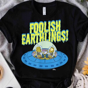 Puede incluir: Una camiseta negra de manga corta con un gráfico de dos alienígenas amarillos con tentáculos dentro de un platillo volante azul. Sobre el platillo, el texto FOOLISH EARTHLINGS! está impreso en una fuente amarilla que gotea con borde azul.