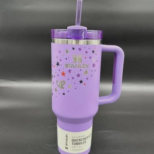 May include: A lavender Stanley Quencher H2.0 Flowstate tumbler with a handle and straw. The tumbler features a star pattern and the Stanley logo. The label reads "QUENCHER H2.0 TUMBLER".