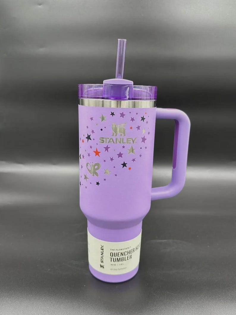 May include: A lavender Stanley Quencher H2.0 Flowstate tumbler with a handle and straw. The tumbler features a star pattern and the Stanley logo. The label reads "QUENCHER H2.0 TUMBLER".