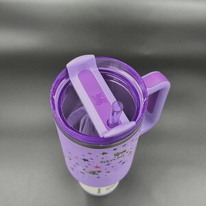 May include: A purple insulated tumbler with a handle and a flip-top lid. The tumbler features a star pattern and the brand name "STANLEY". Includes a clear straw and a purple handle.