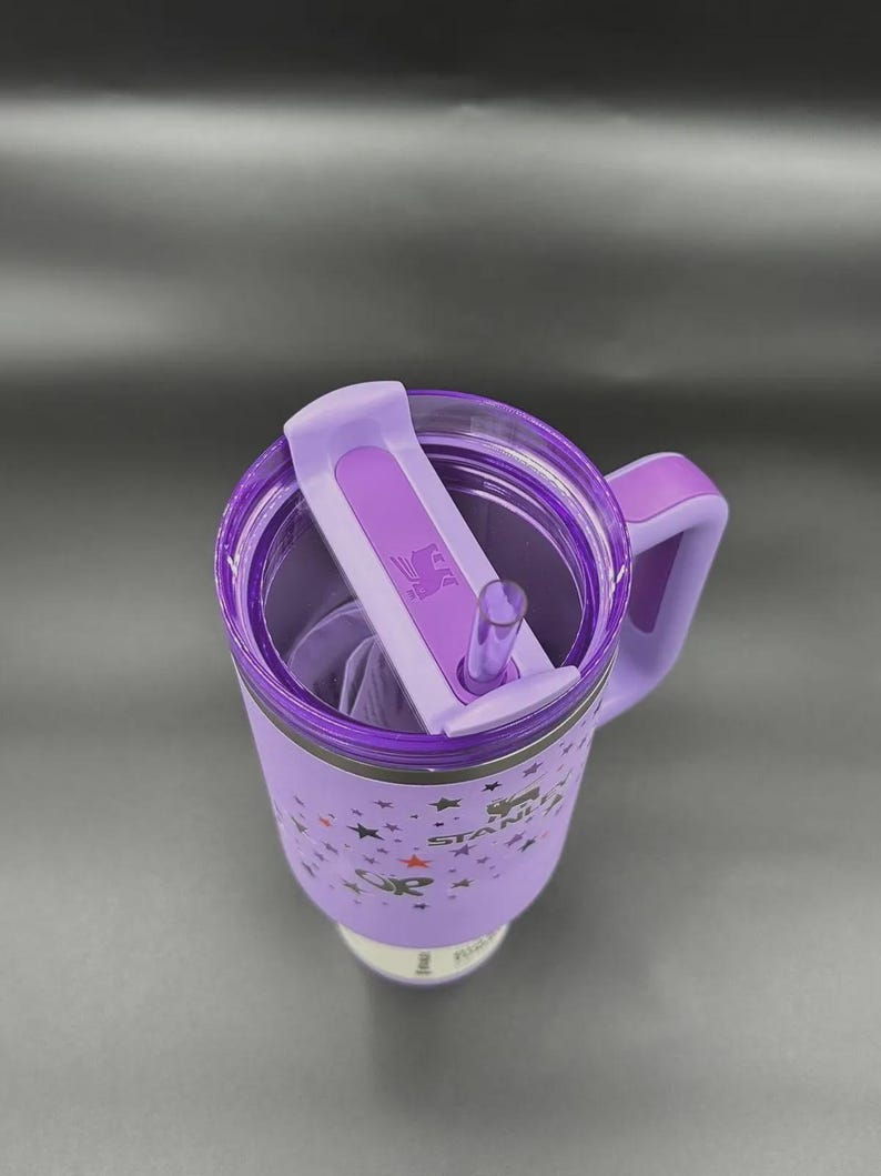 May include: A purple insulated tumbler with a handle and a flip-top lid. The tumbler features a star pattern and the brand name "STANLEY". Includes a clear straw and a purple handle.
