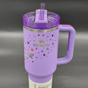 May include: A purple Stanley Quencher H2.0 Flowstate tumbler with a handle and a straw. The tumbler features a star pattern and the Stanley logo. The text "The Flowstate Quencher H2.0 Tumbler" is printed on the bottom.